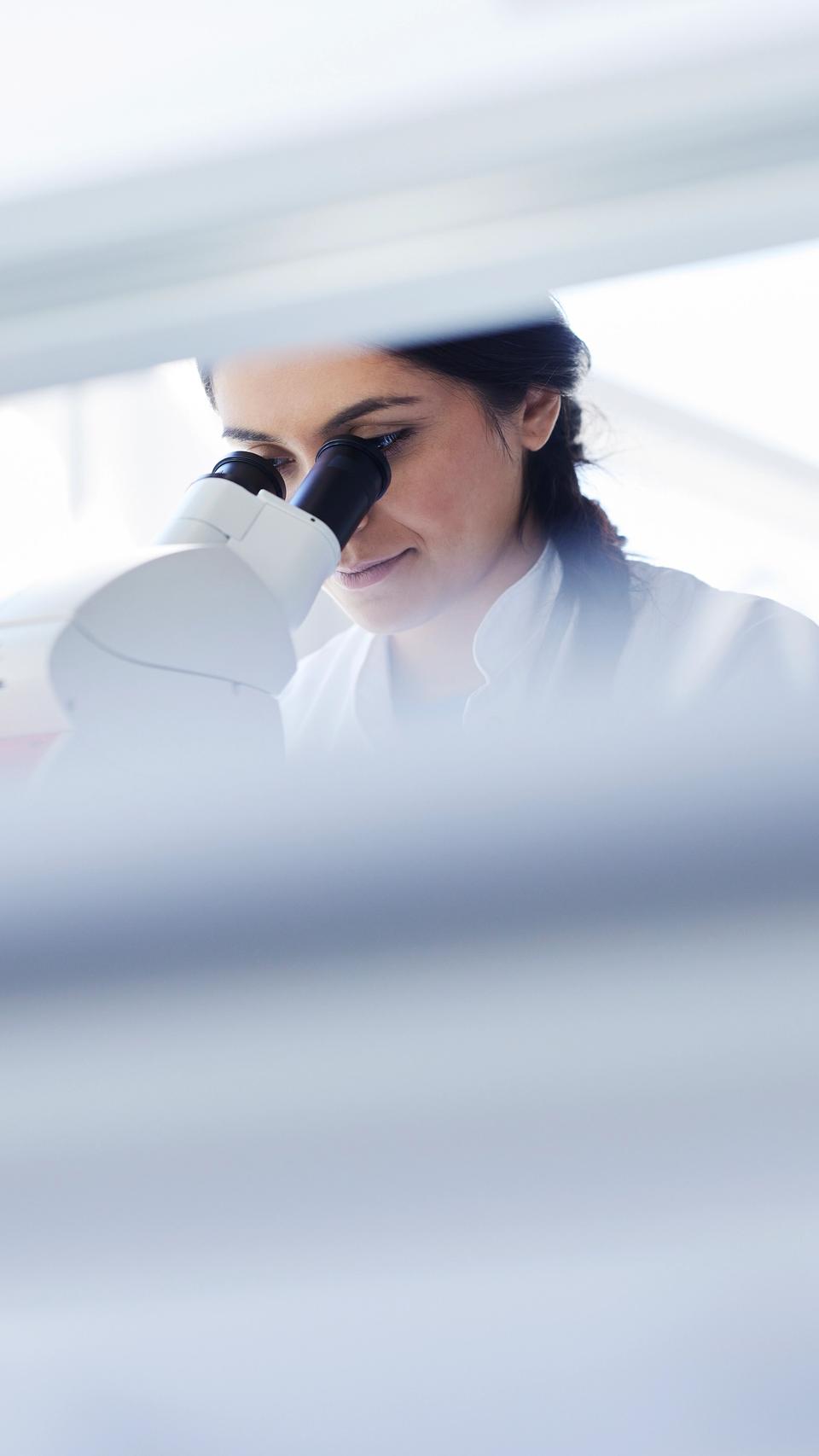 A female scientist looking into a microscope in a brightly lit laboratory.