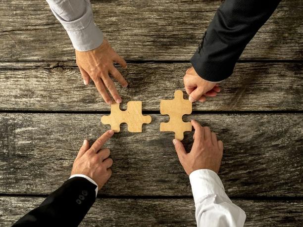 Four hands in business attire are placing two large puzzle pieces together.