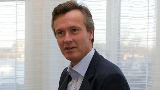 A person in a suit sitting indoors, with sheer curtains and a softly lit background.
