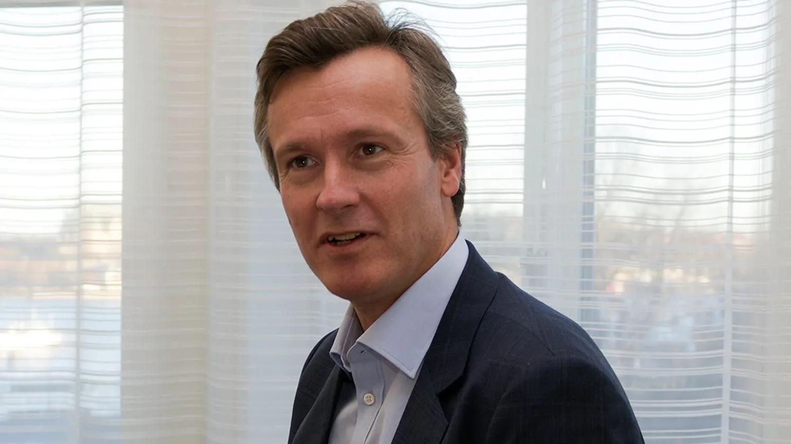 A person in a suit sitting indoors, with sheer curtains and a softly lit background.