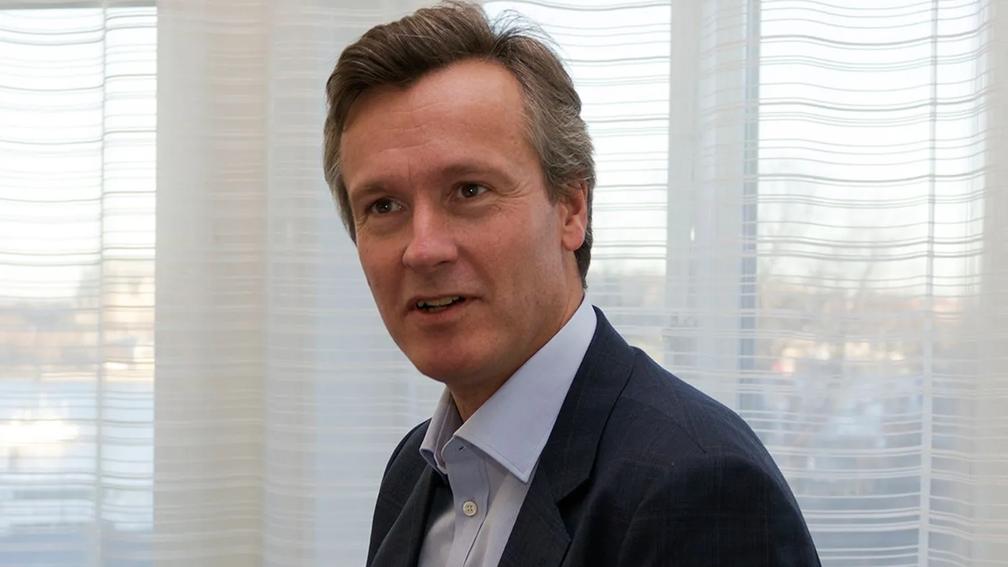 A person in a suit sitting indoors, with sheer curtains and a softly lit background.