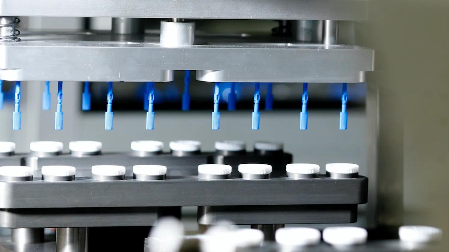 A close-up of a manufacturing machine with blue nozzles above rows of white caps on a conveyor belt.