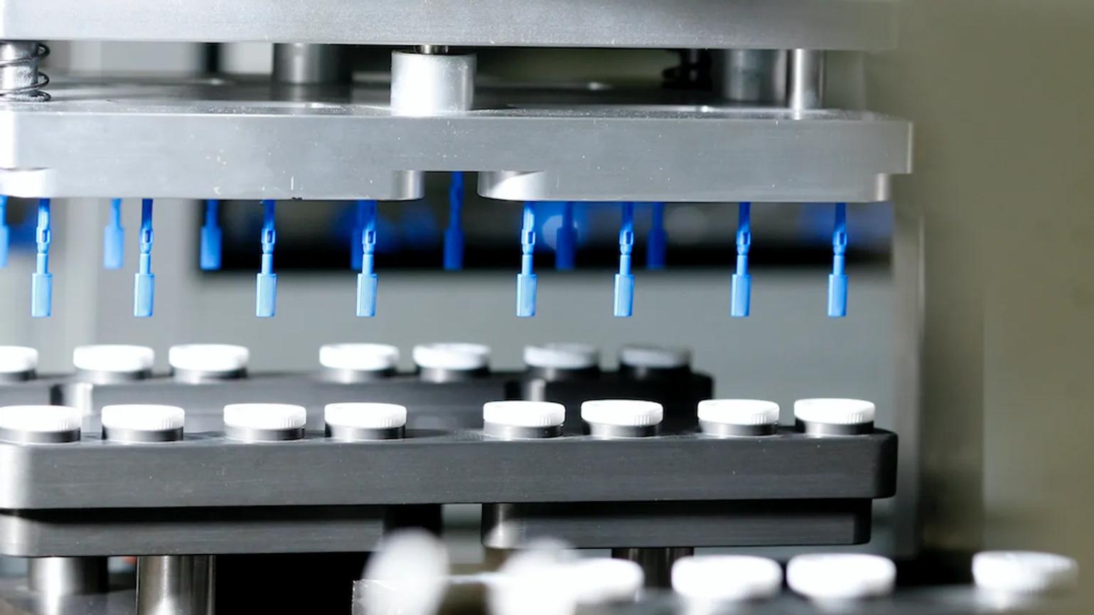 A close-up of a manufacturing machine with blue nozzles above rows of white caps on a conveyor belt.