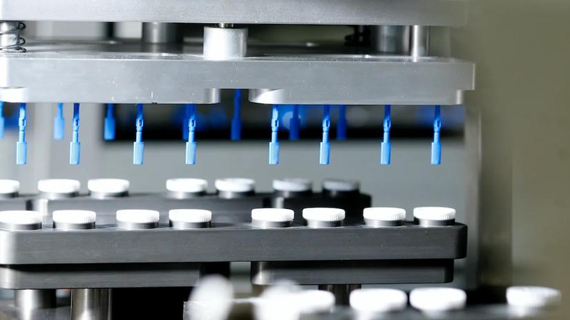 A close-up of a manufacturing machine with blue nozzles above rows of white caps on a conveyor belt.