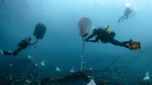 Scuba divers underwater work together to lift a large fishing net using inflatable lift bags, with two more divers in the background holding flashlights.
