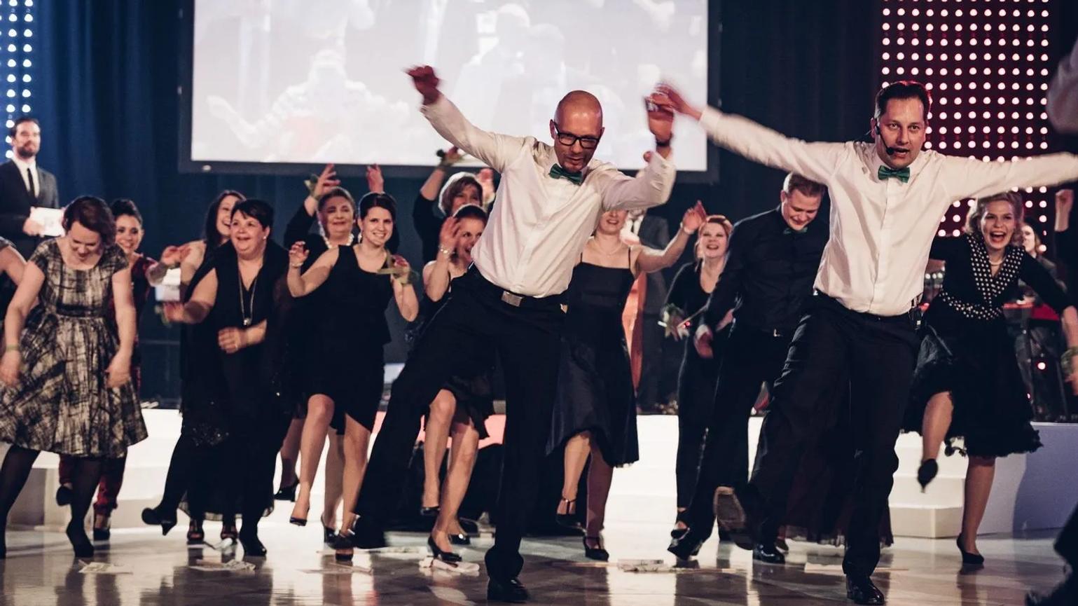 A lively group of people dressed in formal attire, energetically dancing and smiling on a polished dance floor, with a large screen and stage lights in the background.