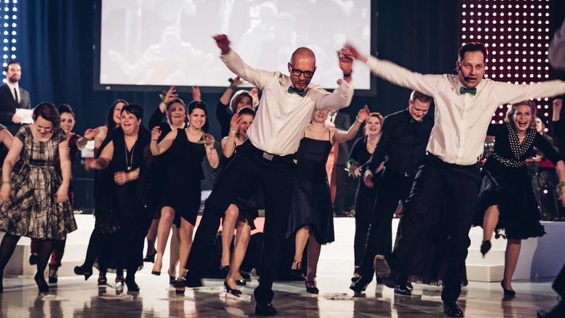 A lively group of people dressed in formal attire, energetically dancing and smiling on a polished dance floor, with a large screen and stage lights in the background.
