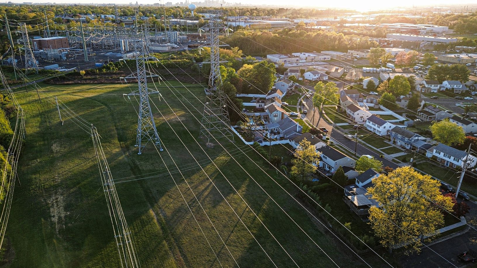 Aerial view of high-voltage power lines and transmission towers running through a suburban neighborhood, with a power substation and trees in the background.