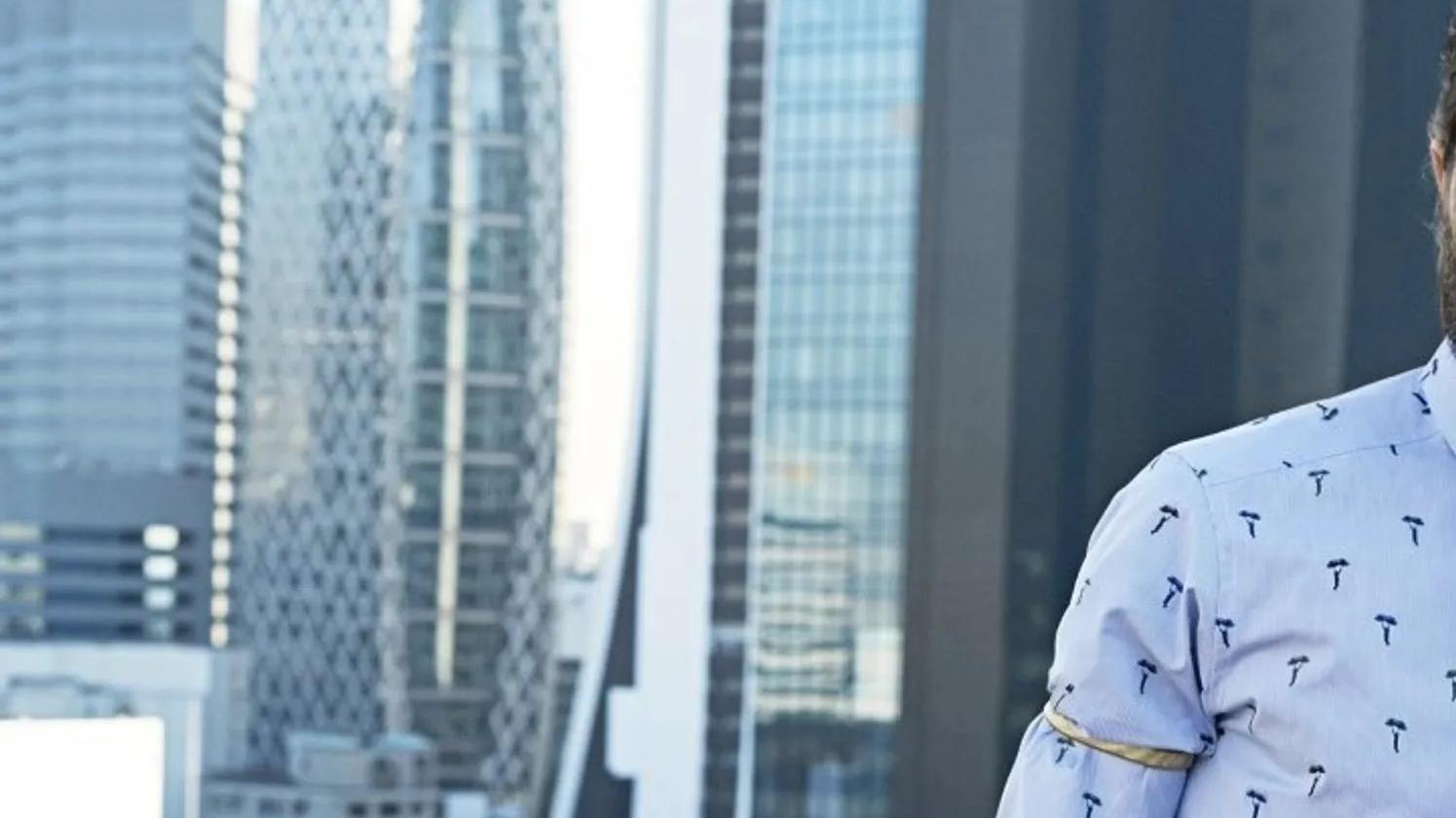 A partial view of a person in a patterned shirt standing in front of a cityscape with modern skyscrapers, including a distinctive building with a geometric facade.