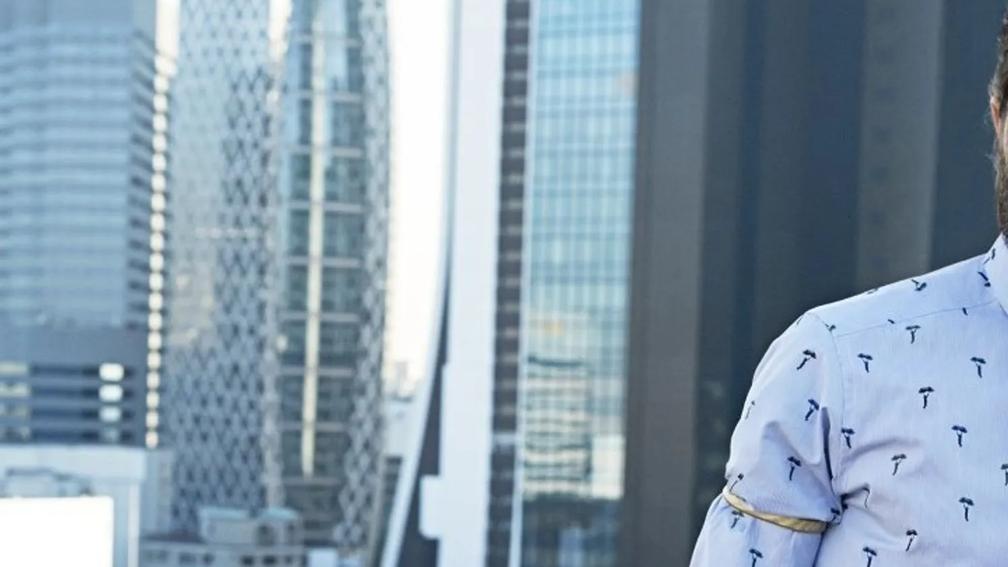 A partial view of a person in a patterned shirt standing in front of a cityscape with modern skyscrapers, including a distinctive building with a geometric facade.