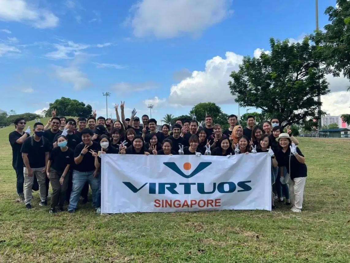 A large group of people outdoors, smiling and posing with peace signs, holding a banner that reads "Virtuos Singapore."