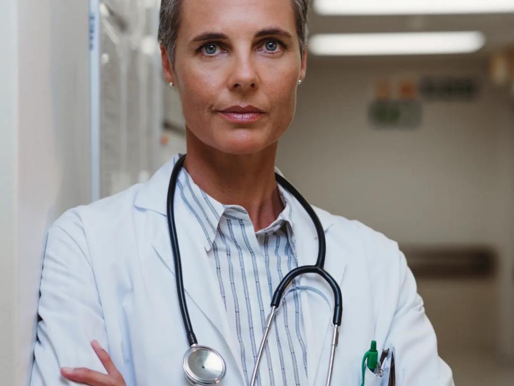 A confident doctor with short hair wearing a white coat and stethoscope, standing in a hospital corridor.