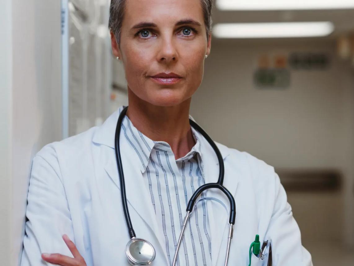 A confident doctor with short hair wearing a white coat and stethoscope, standing in a hospital corridor.