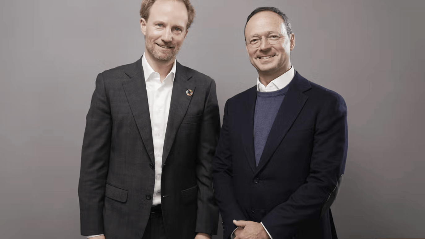 Two men in suits smiling, standing side by side against a plain background.