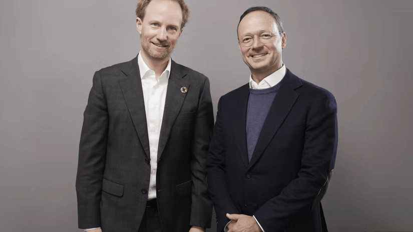 Two men in suits smiling, standing side by side against a plain background.