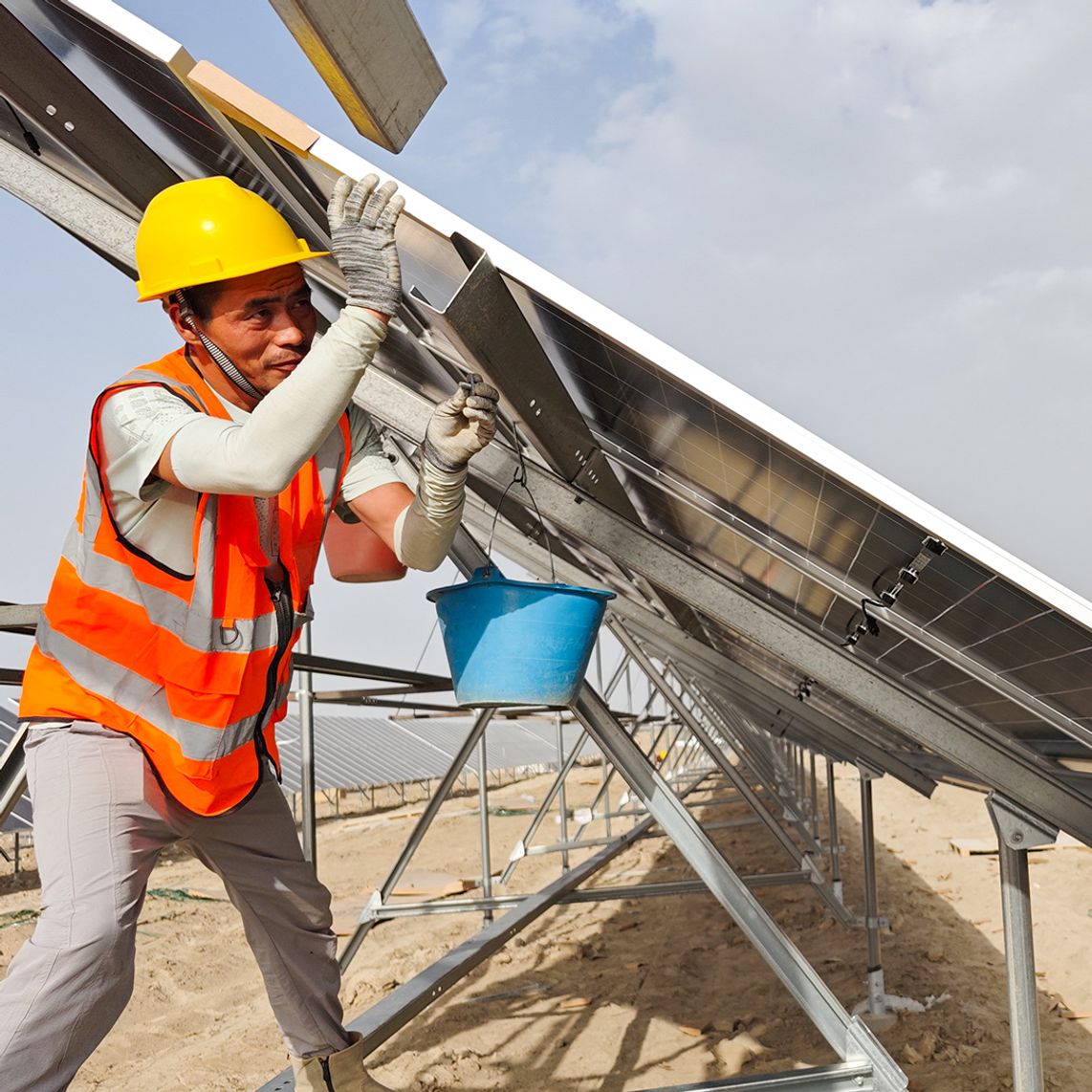 Two workers in orange safety vests and yellow helmets install a solar panel on a metal frame at a solar farm construction site.