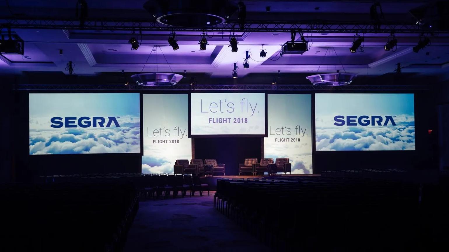 Stage setup for a conference with large screens displaying "Let's fly. FLIGHT 2018" and "SEGRA" above a cloud background, with seating arranged in front.