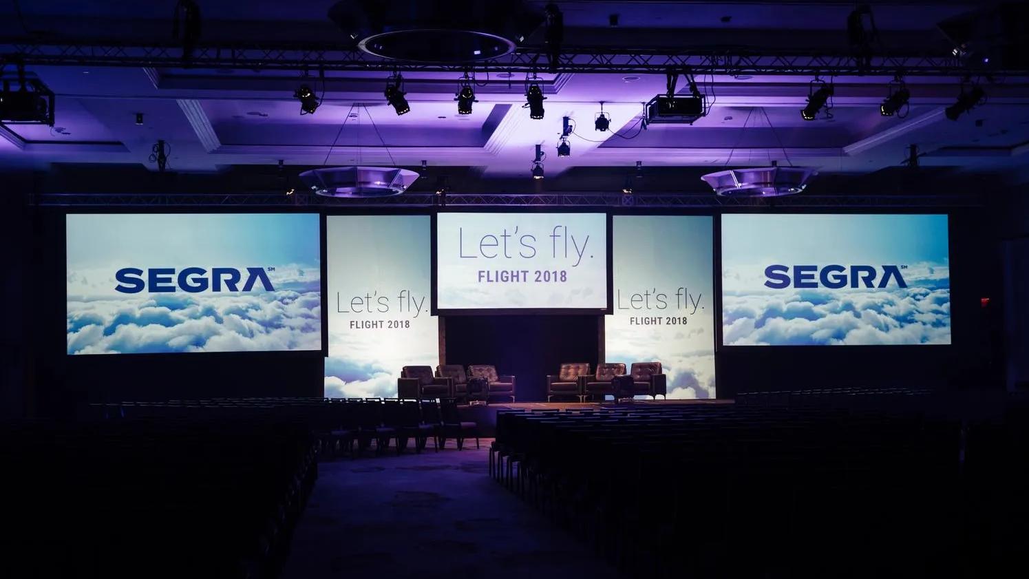 Stage setup for a conference with large screens displaying "Let's fly. FLIGHT 2018" and "SEGRA" above a cloud background, with seating arranged in front.