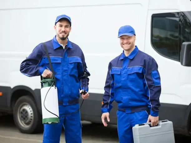 Two smiling workers in blue uniforms and caps stand in front of a white van.