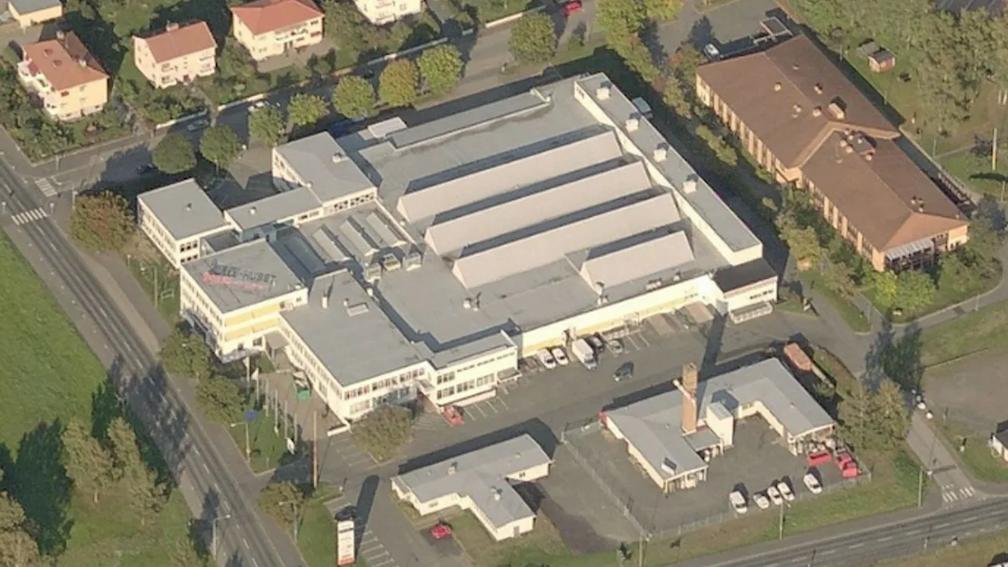Aerial view of a large industrial building with a white roof, surrounded by trees and residential houses.