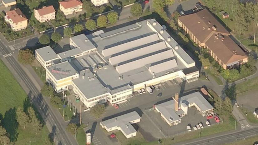 Aerial view of a large industrial building with a white roof, surrounded by trees and residential houses.