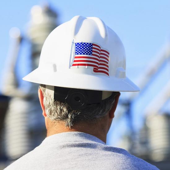A worker wearing a white hard hat with an American flag sticker stands facing an industrial facility, seen from behind.