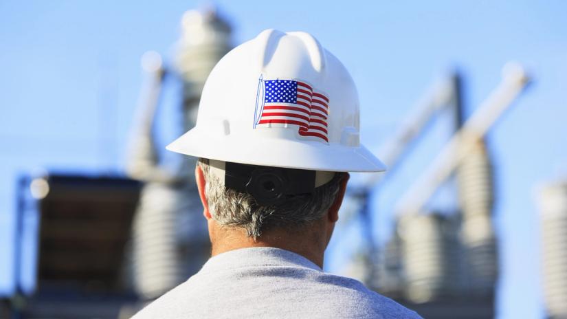A worker wearing a white hard hat with an American flag sticker stands facing an industrial facility, seen from behind.