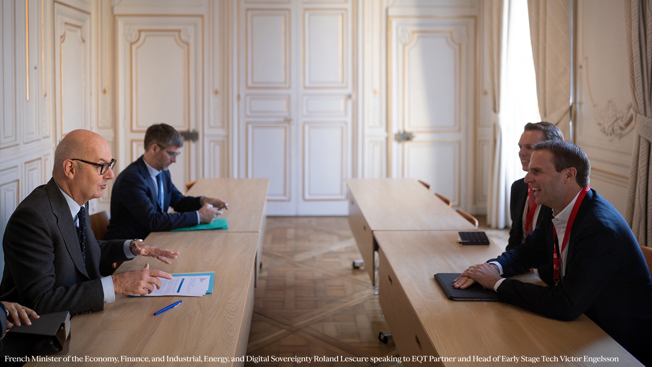 A group of business professionals sitting across long wooden tables in an ornate conference room, engaged in a discussion.