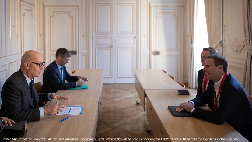 A group of business professionals sitting across long wooden tables in an ornate conference room, engaged in a discussion.