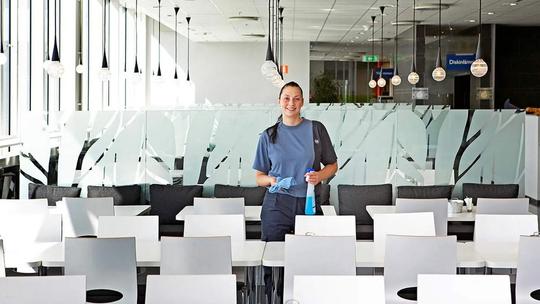 A woman in a blue uniform holding cleaning supplies, standing in a bright cafeteria with white tables and chairs.