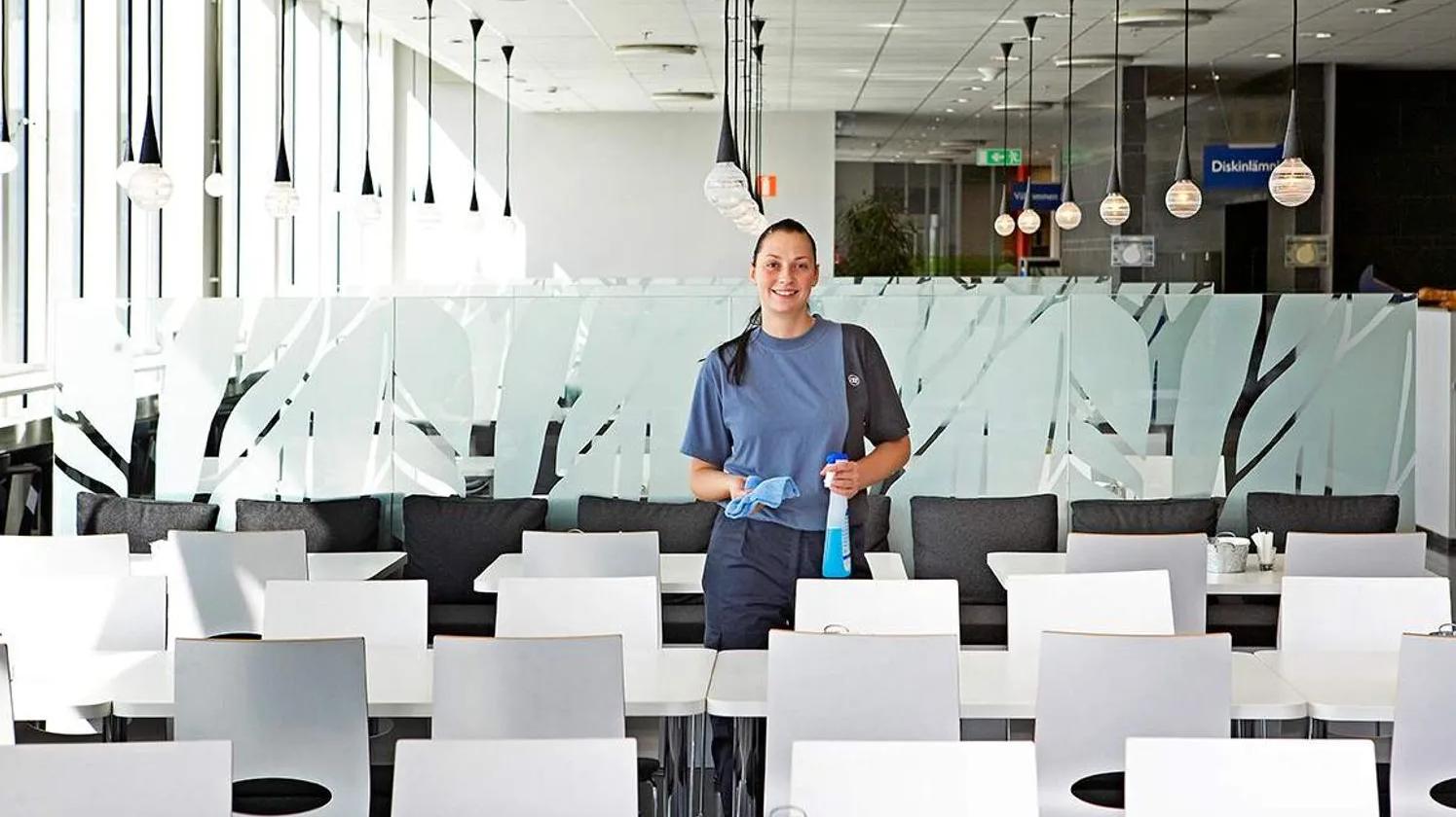 A woman in a blue uniform holding cleaning supplies, standing in a bright cafeteria with white tables and chairs.
