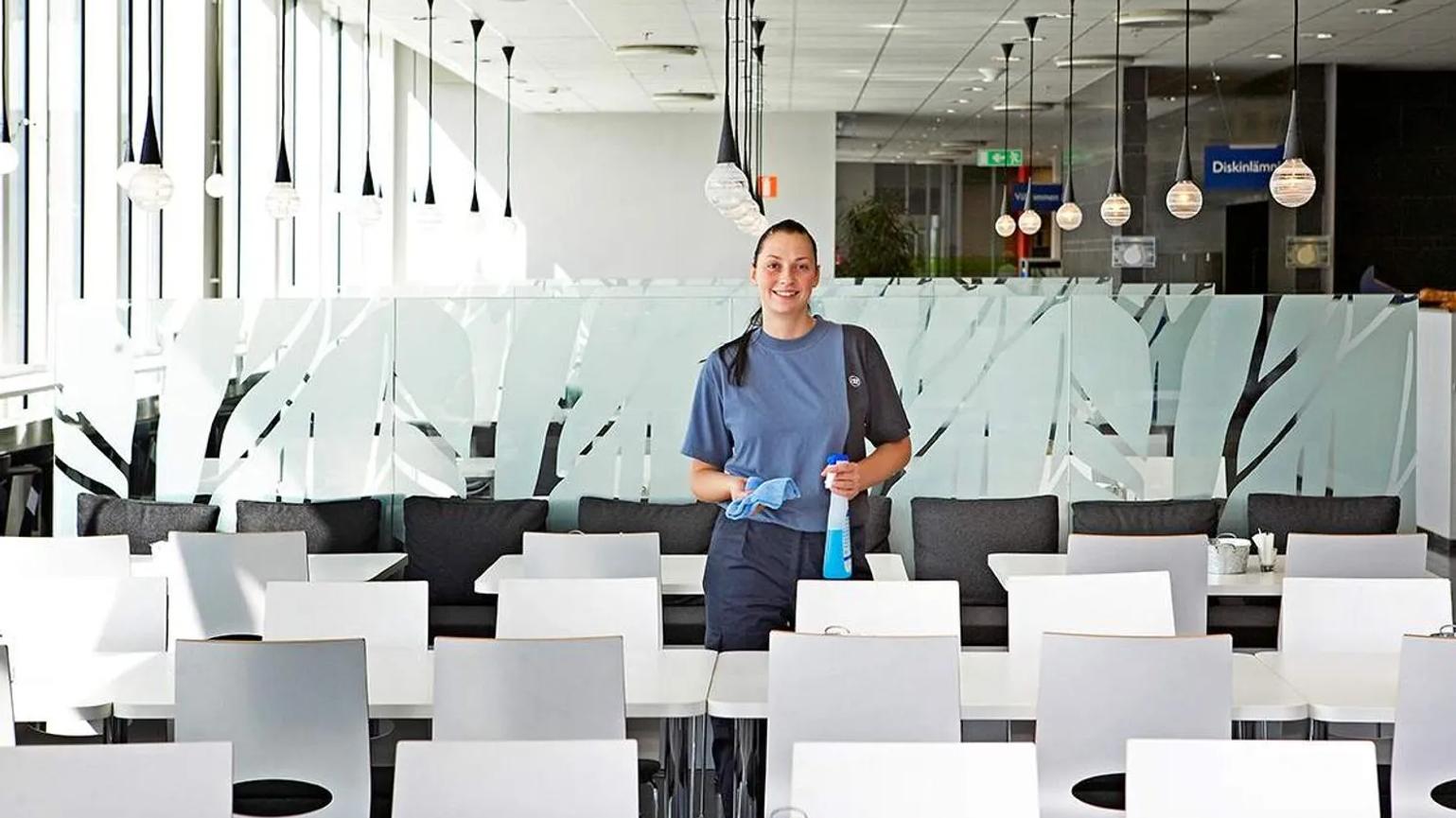 A woman in a blue uniform holding cleaning supplies, standing in a bright cafeteria with white tables and chairs.