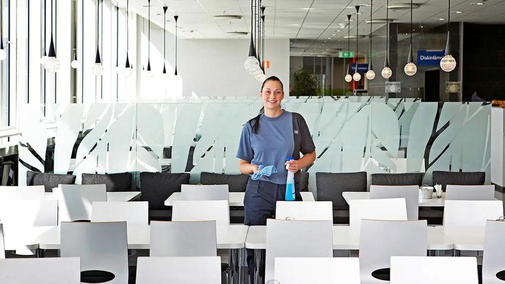 A woman in a blue uniform holding cleaning supplies, standing in a bright cafeteria with white tables and chairs.