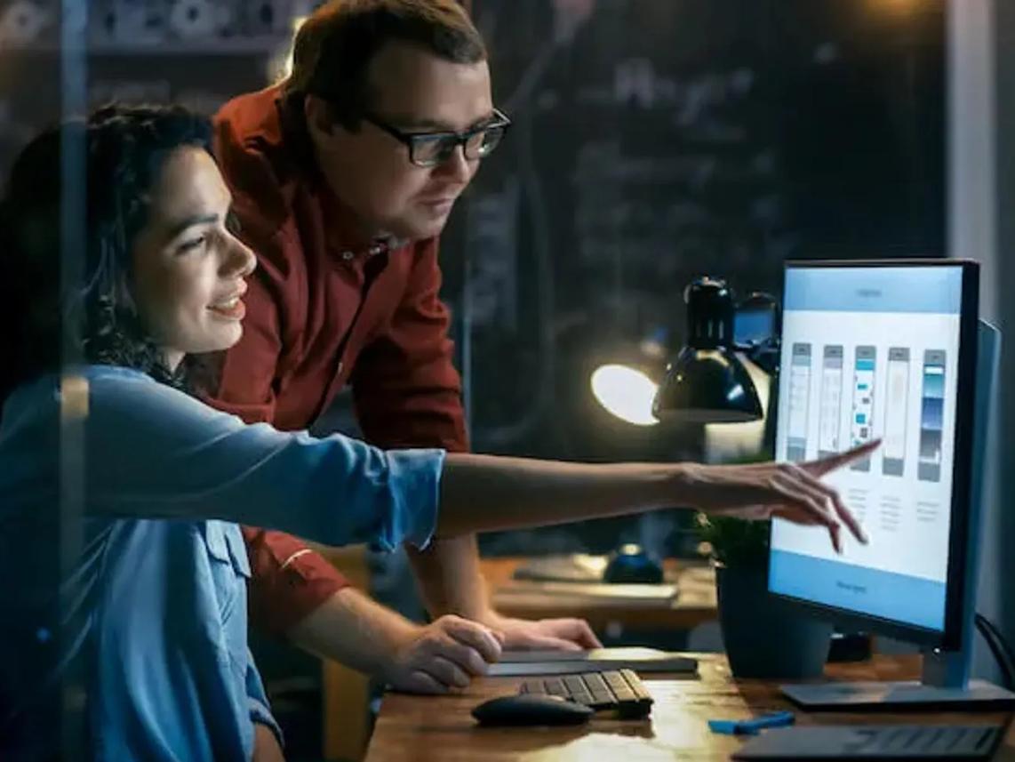 Two colleagues working together at a desk, with one pointing at a computer screen displaying a design interface
