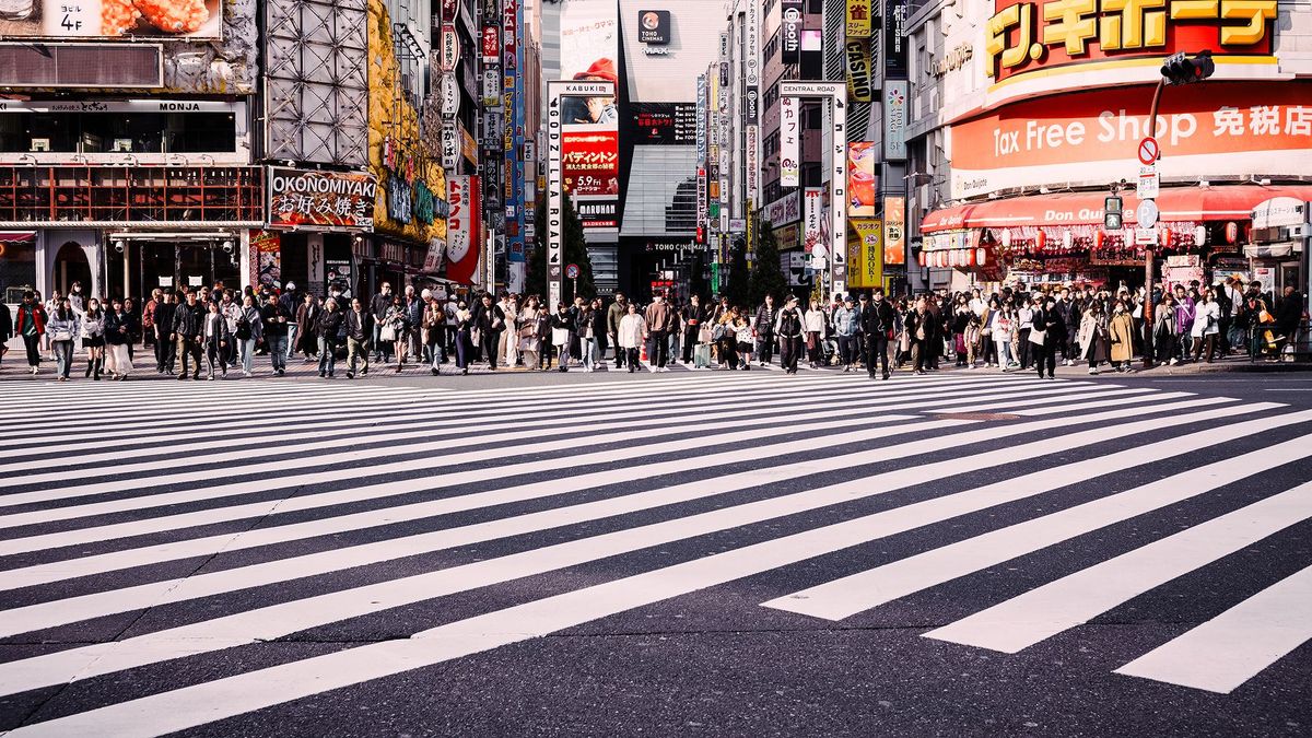 Large crowd waiting to cross a wide zebra crossing in a busy city street lined with bright shop signs.
