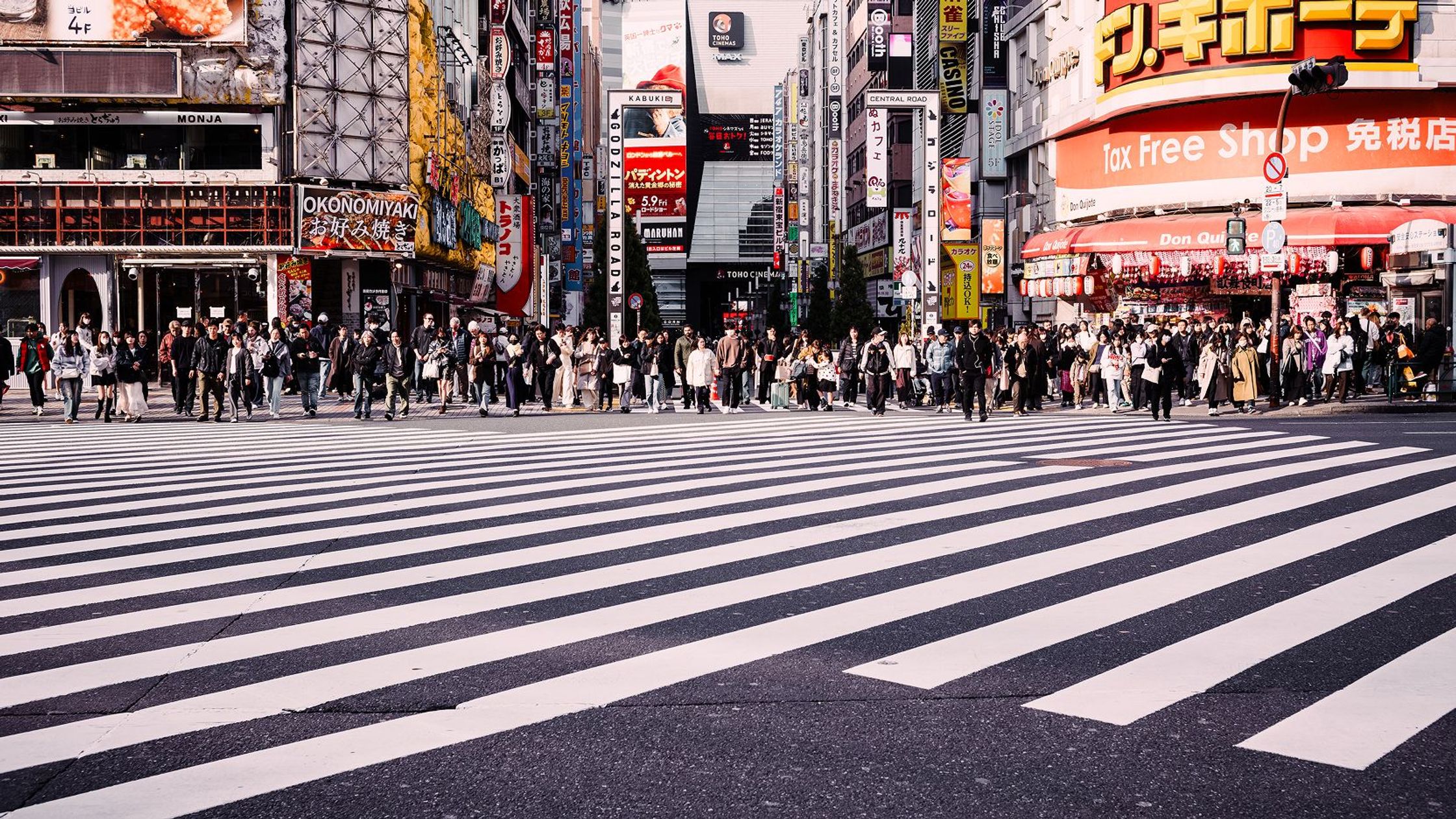 Large crowd waiting to cross a wide zebra crossing in a busy city street lined with bright shop signs.