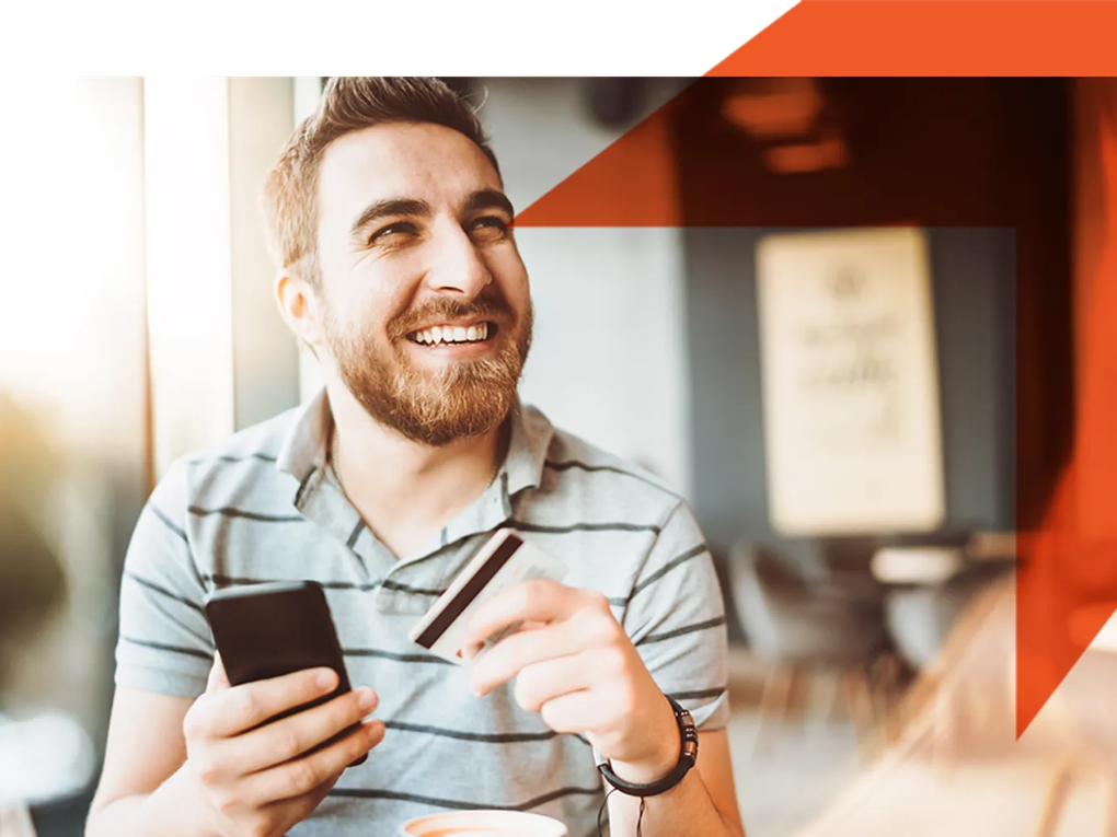 A smiling man holding a smartphone and a credit card, sitting in a bright cafe.