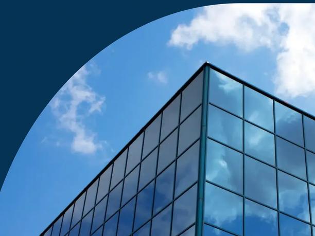 A modern glass building corner reflecting the sky and clouds, with a dark blue curved overlay at the top.