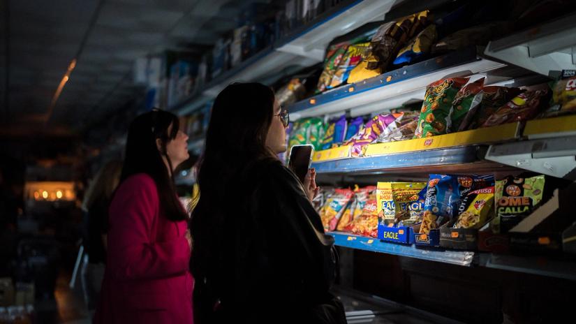 Two women use a phone as a flashlight to shop for snacks in a dark store during a power outage.