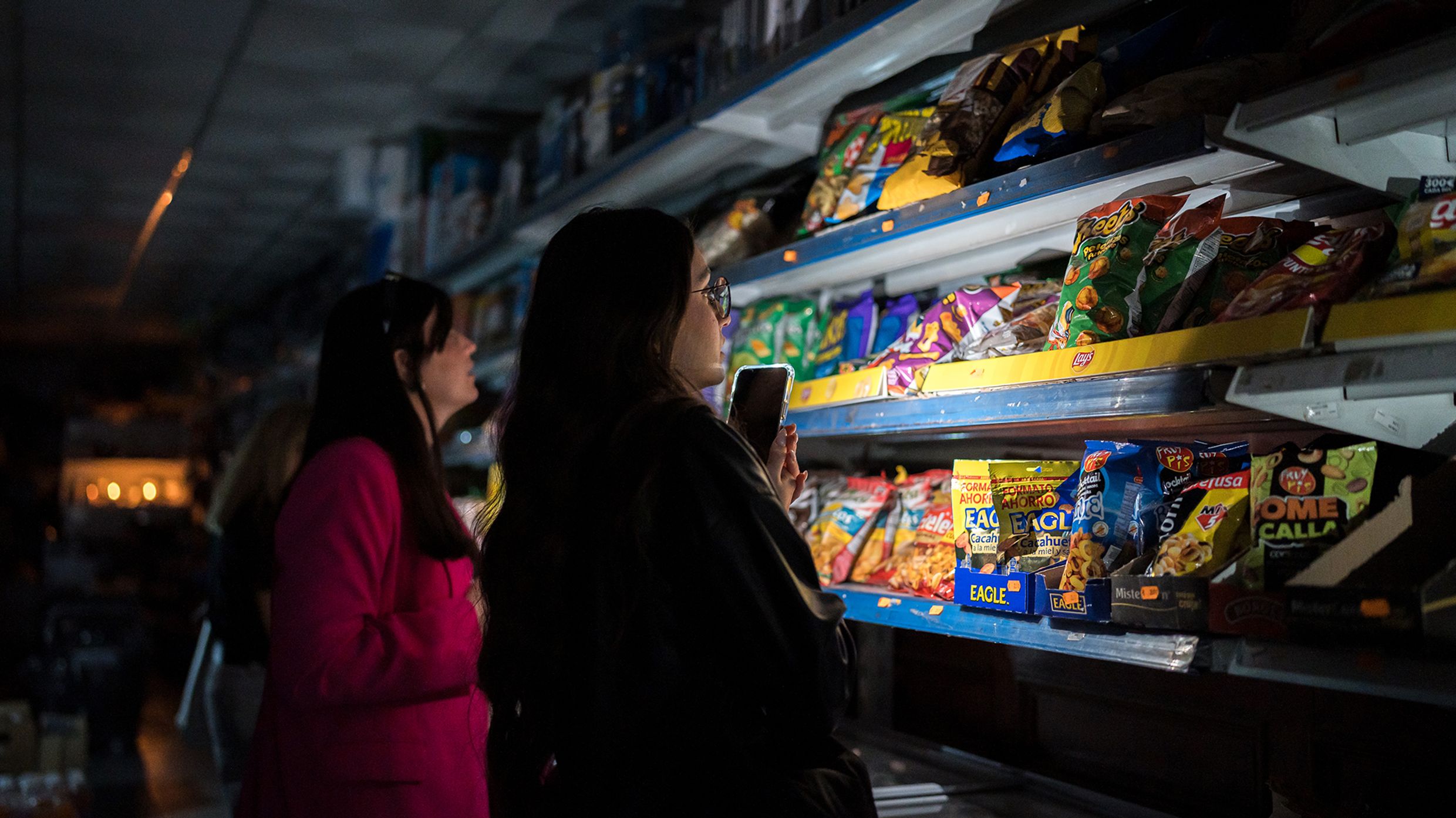 Two women use a phone as a flashlight to shop for snacks in a dark store during a power outage.
