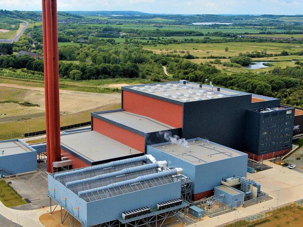 A large industrial facility with multiple buildings and tall chimneys, set in a rural landscape with fields and trees.