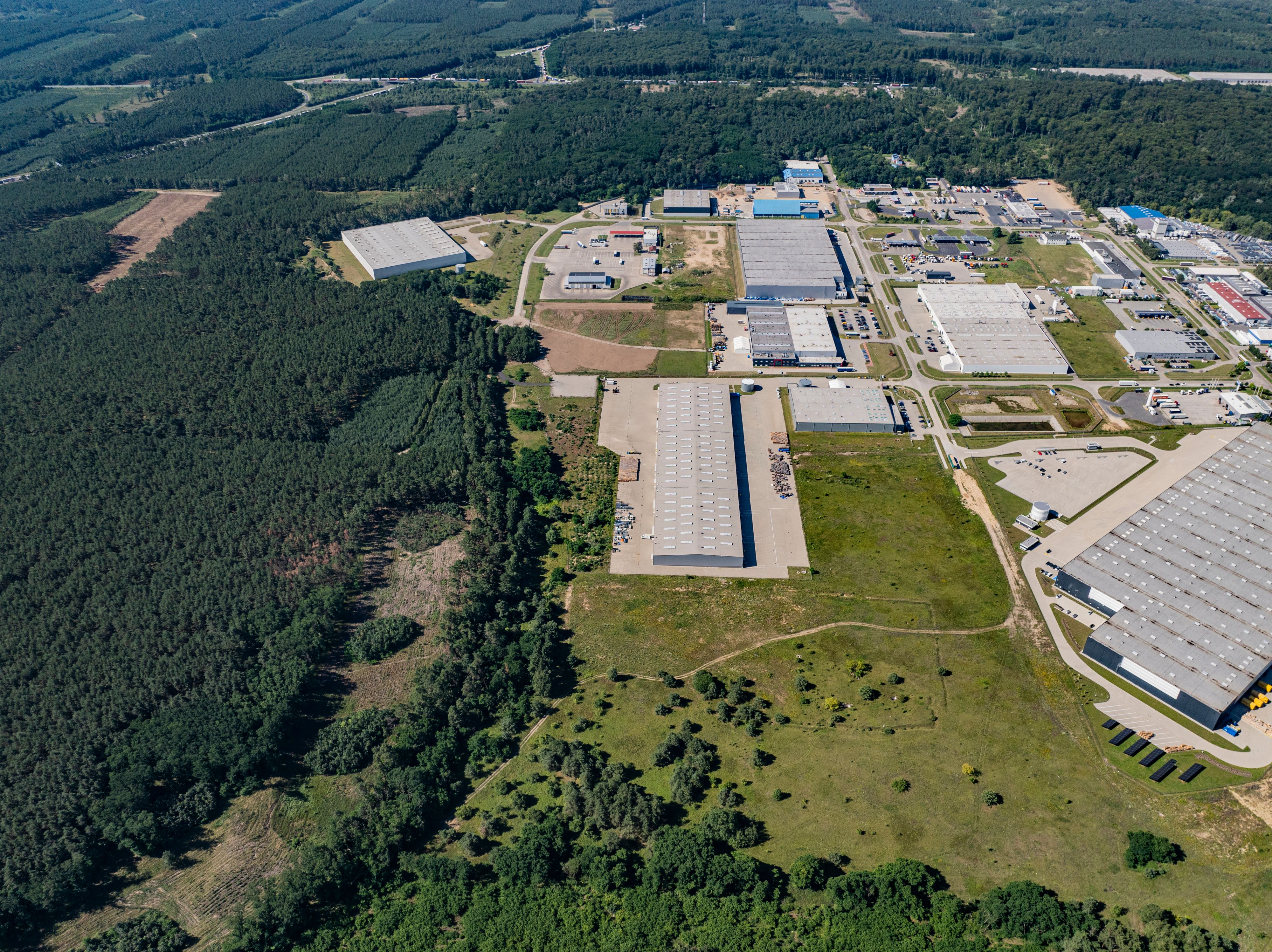 Aerial view showing warehouses and industrial buildings next to a dense forest, with roads and green spaces separating the structures.