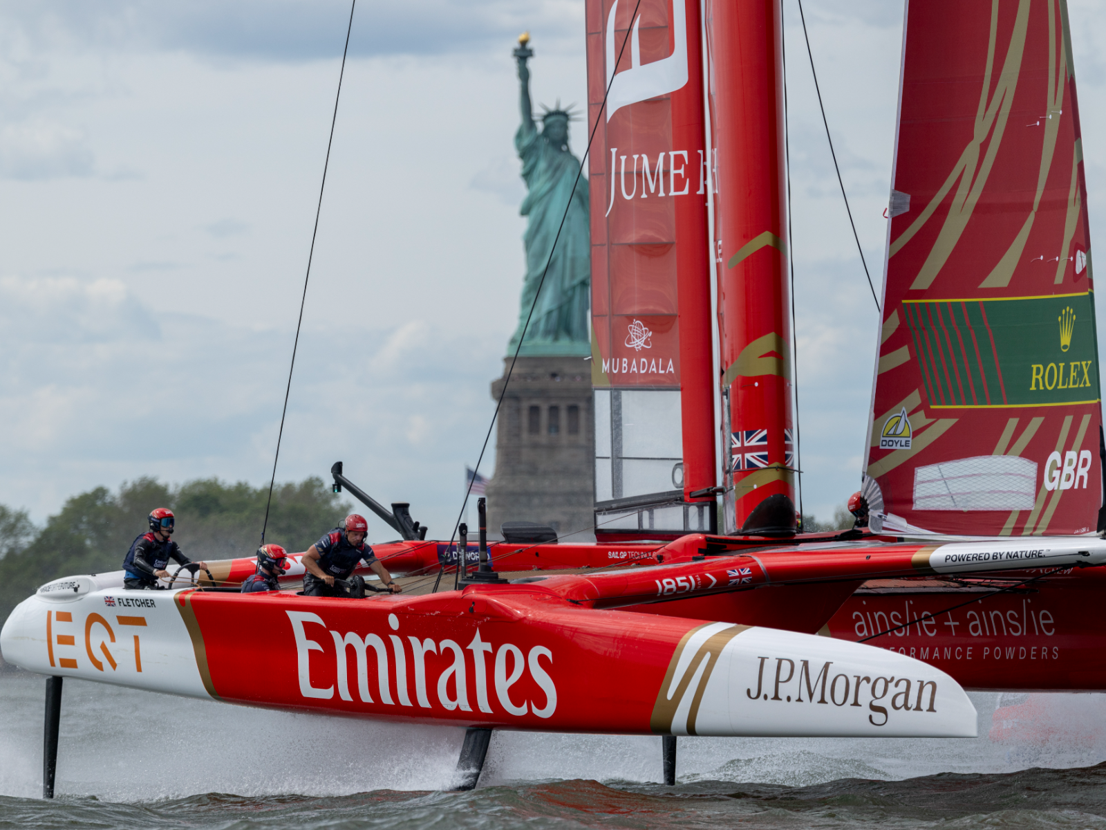 A red and white Emirates Great Britain catamaran race boat foiling across the water in front of the Statue of Liberty in New York Harbor.