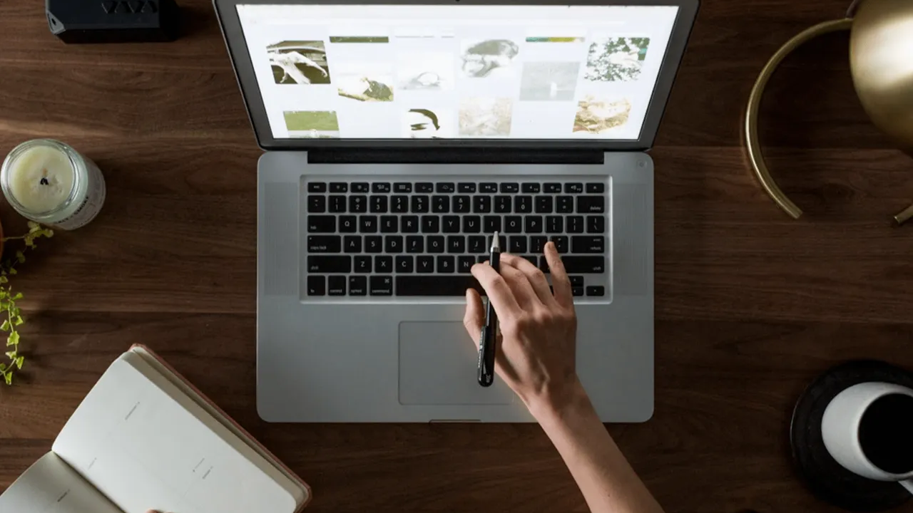 A person using a laptop on a wooden desk, browsing images, with a notebook, candle, and coffee cup nearby.