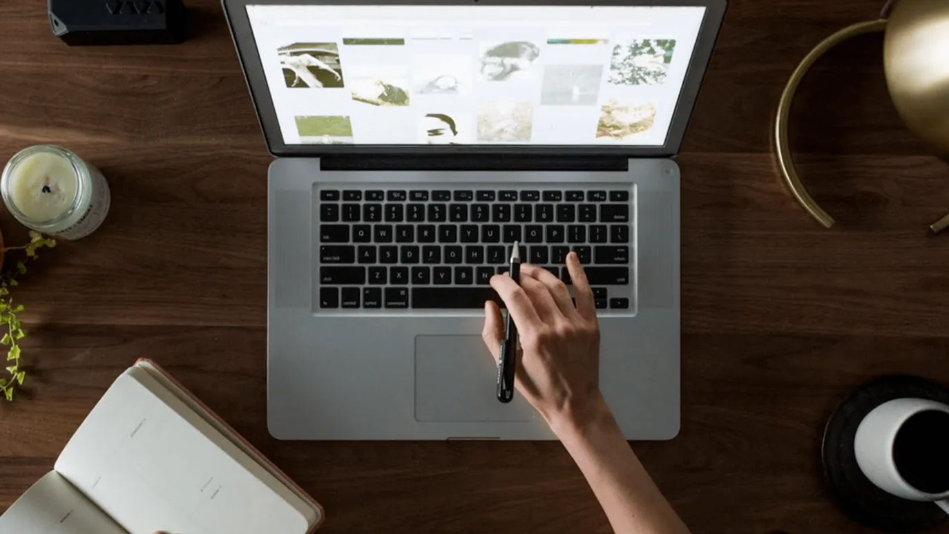 A person using a laptop on a wooden desk, browsing images, with a notebook, candle, and coffee cup nearby.