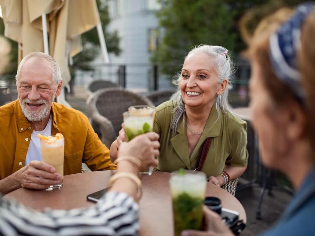 Elderly friends sitting outdoors at a table, smiling and clinking glasses filled with colorful drinks.