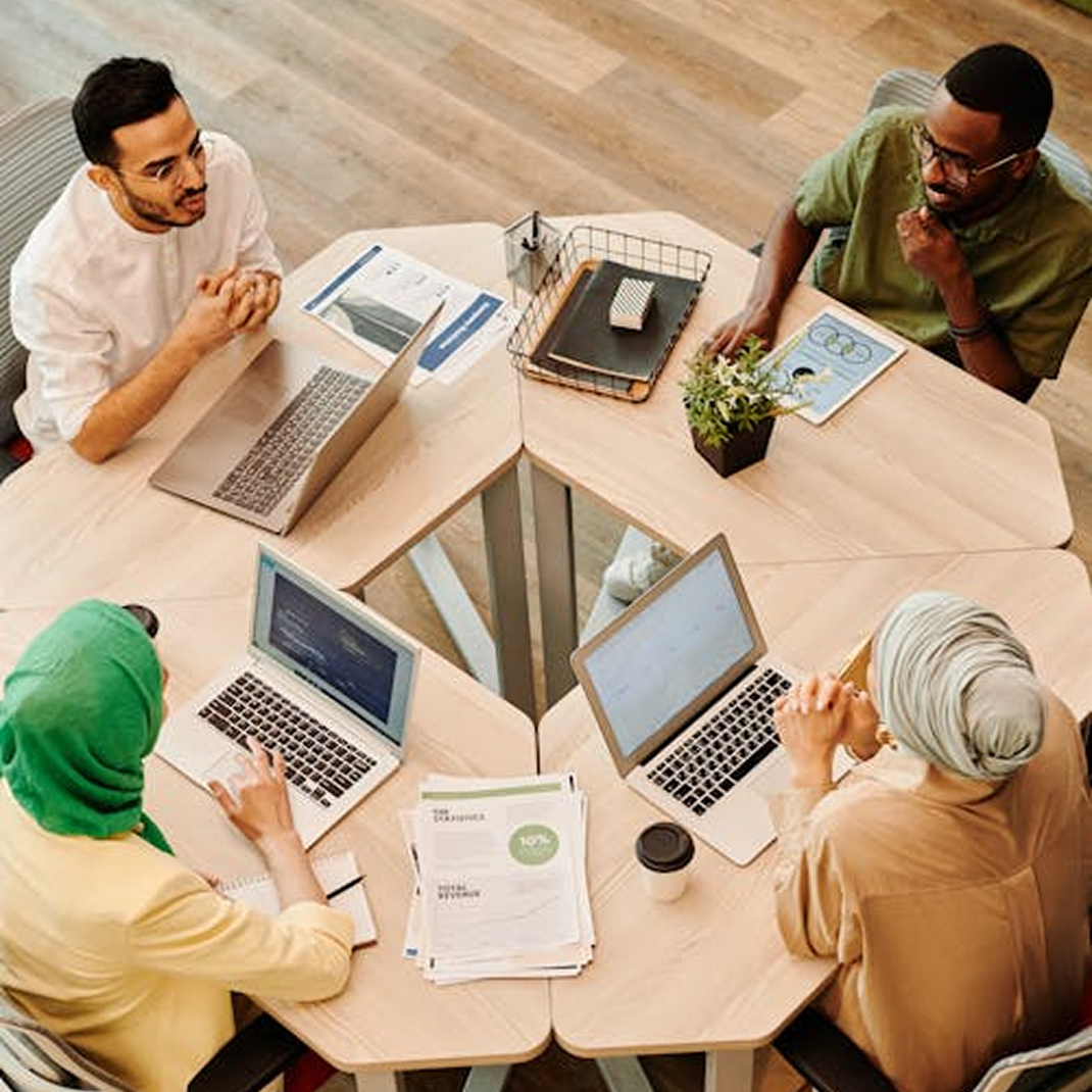 An overhead shot of four colleagues in business casual attire sitting around a modular wooden desk configuration, collaborating with laptops, tablets, and paperwork.