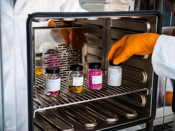 A gloved hand placing a jar with a white substance into a laboratory oven.