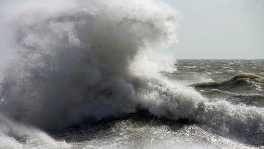 A powerful ocean wave crashing with force, creating a large spray of white foam against a stormy sea backdrop.