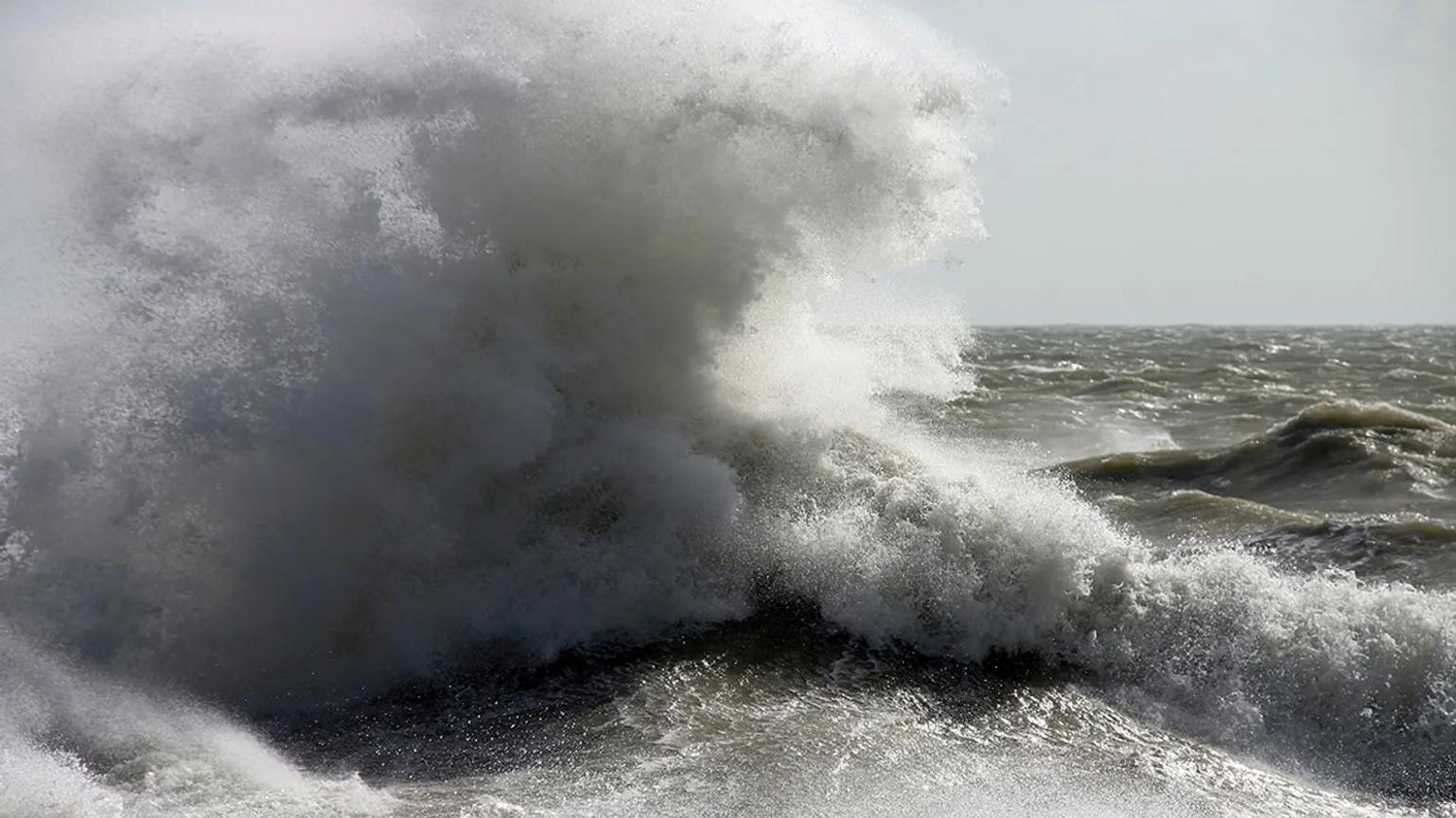 A powerful ocean wave crashing with force, creating a large spray of white foam against a stormy sea backdrop.